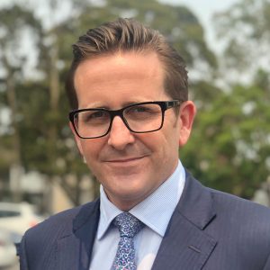 A man wearing a grey and blue suit with black-framed glasses poses outside.