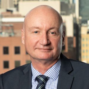 A man wearing a grey suit seated at a desk, city skyline visible behind him.