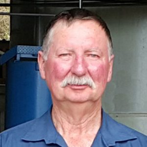 A man with a grey moustache in a blue collared shirt stands in front of some industrial machinery.