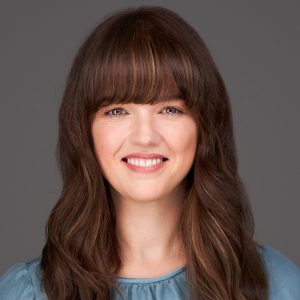 A woman with long brown hair and a steel-blue top in front of a dark grey background.